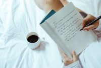 Seorang wanita menikmati momen tenang sembari menulis catatan harian di atas buku agenda kayu dengan secangkir kopi hangat di sampingnya. Kebiasaan menulis ini membantu menenangkan pikiran dan meredakan stres harian secara efektif.