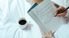 Seorang wanita menikmati momen tenang sembari menulis catatan harian di atas buku agenda kayu dengan secangkir kopi hangat di sampingnya. Kebiasaan menulis ini membantu menenangkan pikiran dan meredakan stres harian secara efektif.